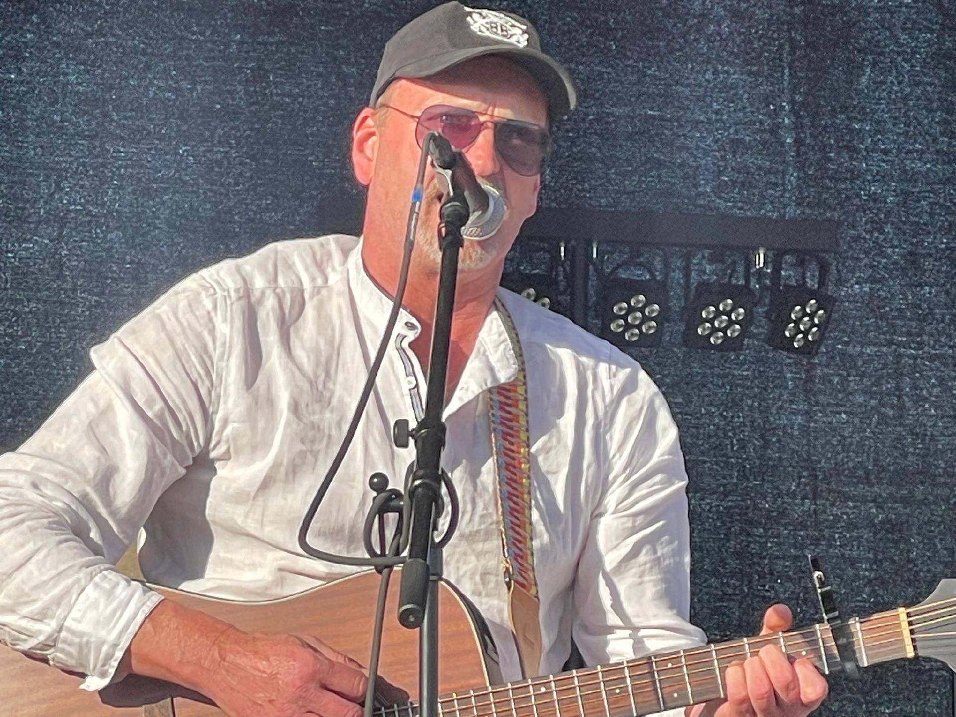 Andy steht mit seiner akustischen Gitarre vor einem Mikrofon auf der Bühne. Er trägt eine Baseballkappe, Sonnenbrille und ein weißes Hemd