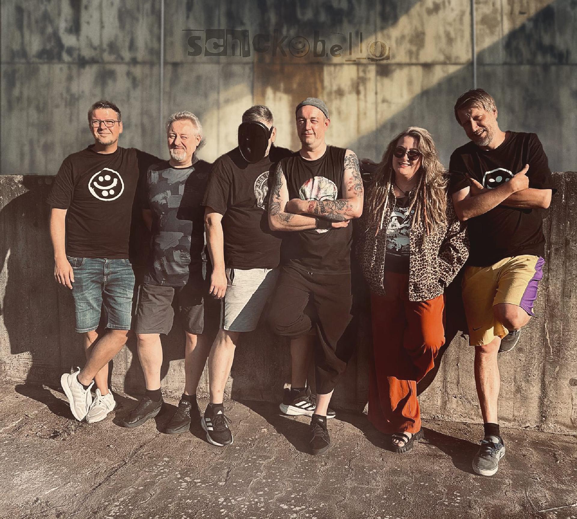 Sechs Personen posieren fröhlich nebeneinander an einer Betonwand, sommerlich gekleidet bei Sonnenlicht.